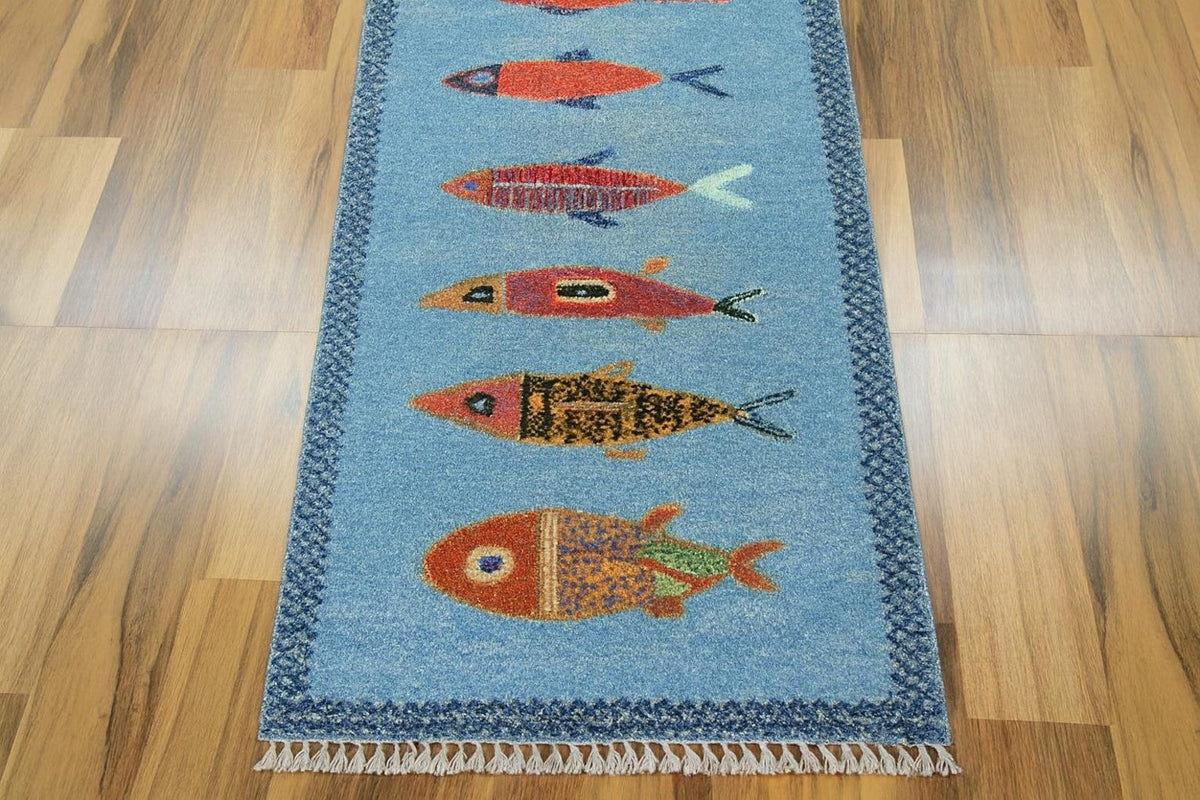 A rectangular blue rug with a fringe bottom edge, featuring a vertical row of stylized fish in orange, red, yellow, and green, laid out on a light brown wooden floor.