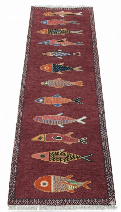 Full view of handmade fish runner rug featuring colorful fish designs on a deep red background with fringe edges.