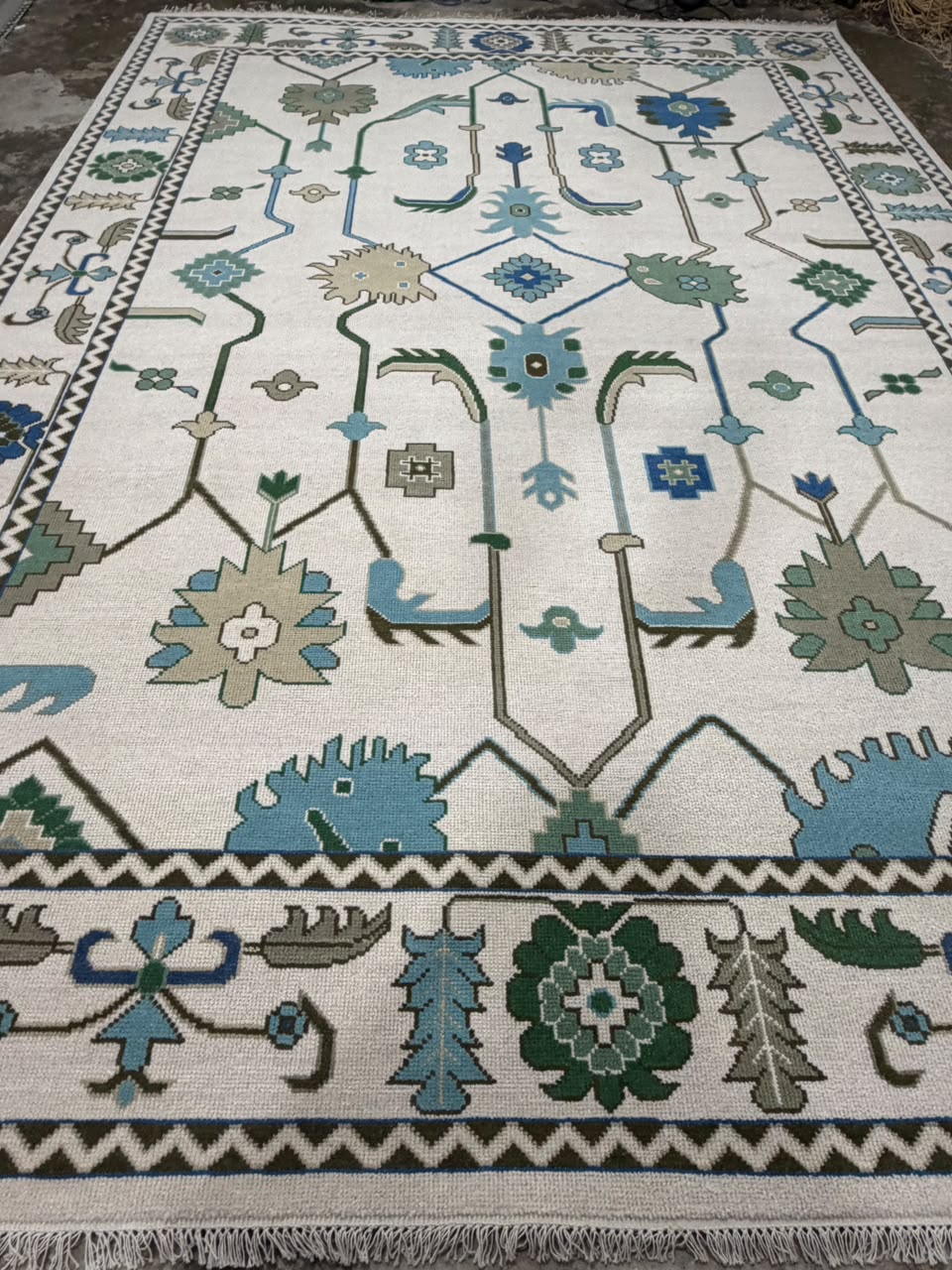 Ivory area rug with blue and green floral geometric motifs, zigzag border and fringed ends.