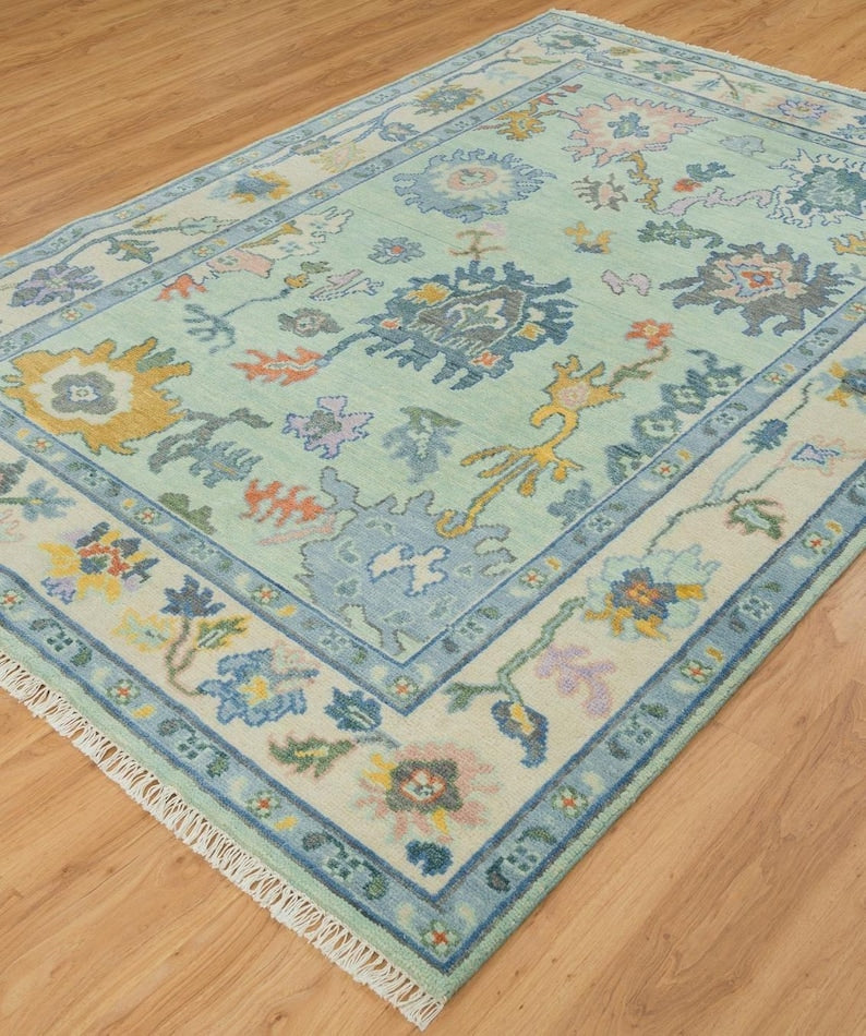 Angled view of a mint green floral Oushak rug with blue outlines, an ivory border and cream fringe