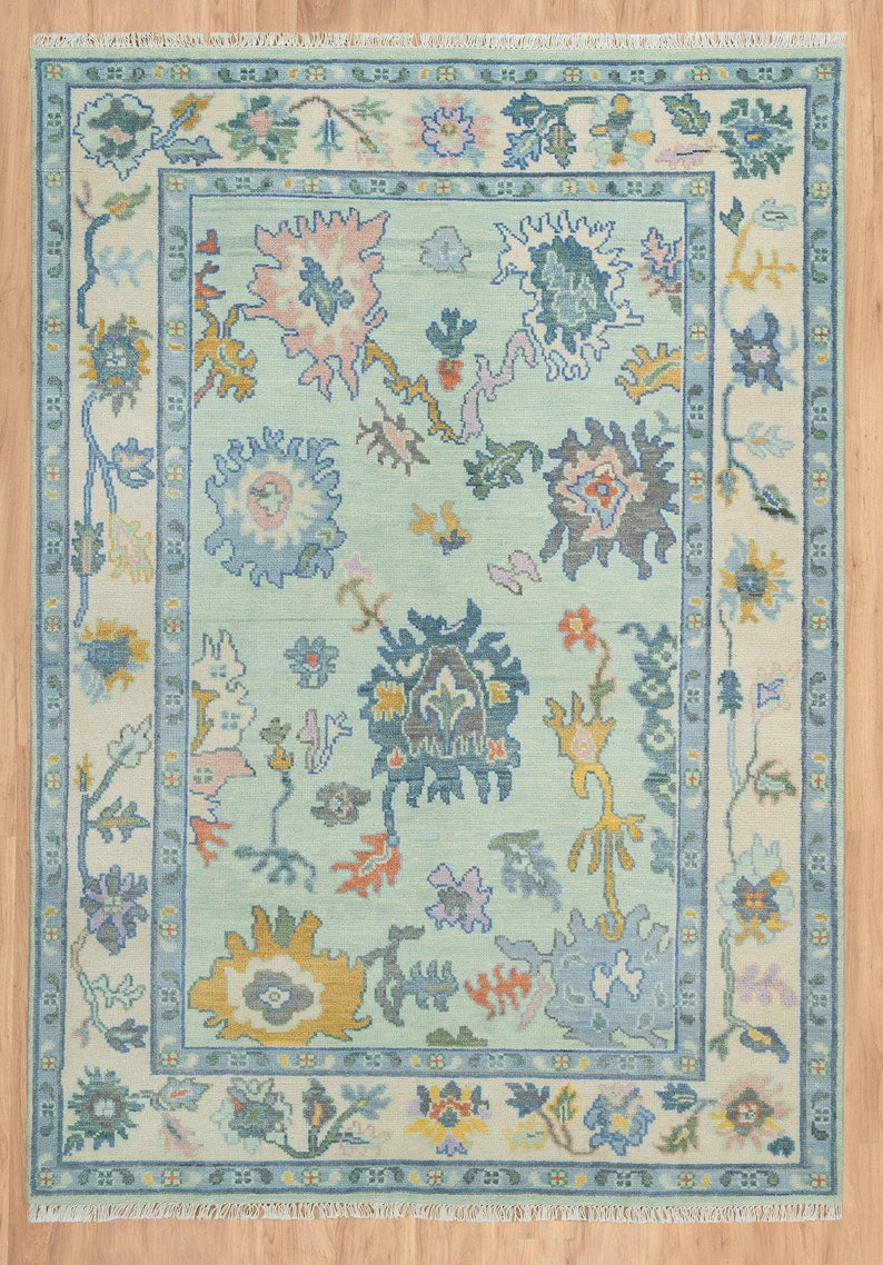 Full view of a mint green floral Oushak rug with pastel motifs, an ivory border and cream fringe.